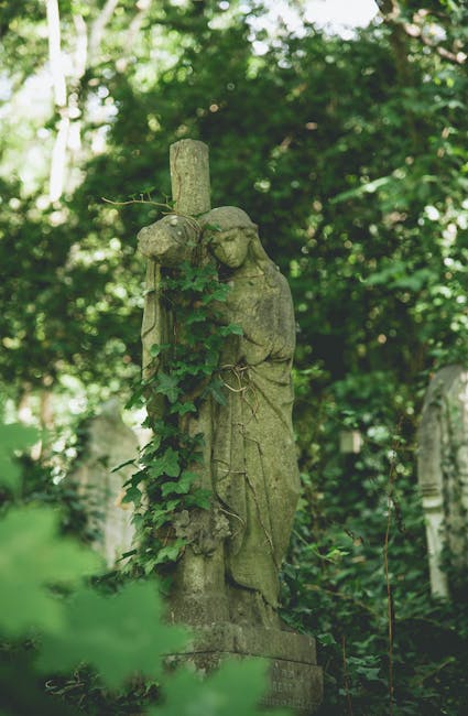 A weathered stone statue of an angel with closed eyes and clasped hands, standing amidst lush green foliage in a garden or park setting. The angel's head is slightly bowed, and the surface of the scul