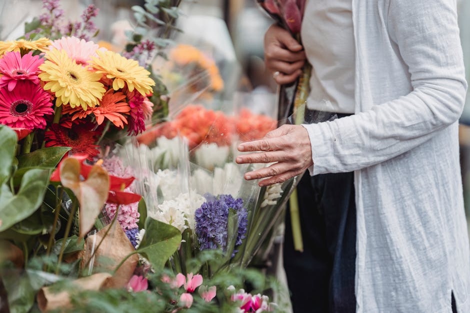 A person in a light grey cardigan and dark trousers is reaching out with their right hand toward a display of fresh flowers at a market or flower shop. The bouquet features vibrant pink, yellow, white