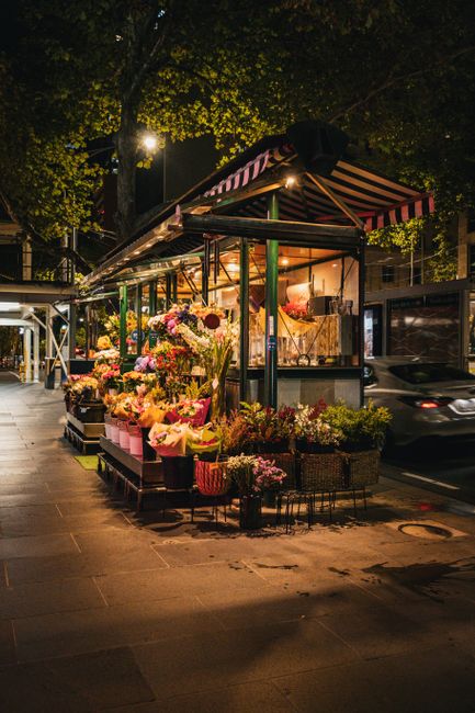 A flower stall illuminated by warm artificial lighting at night, displaying an arrangement of fresh flowers including pink roses, white lilies, and purple chrysanthemums, wrapped in pastel-colored pap