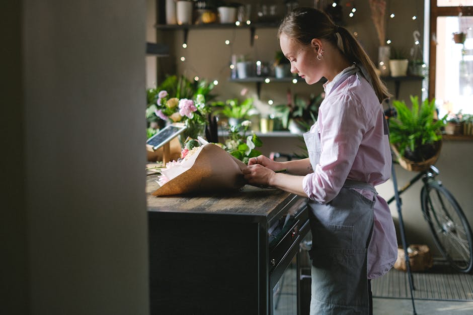 A young female florist in a light pink shirt and gray apron arranging a bouquet of fresh flowers on a dark wooden countertop in a cozy, well-lit floral shop. The bouquet includes pink roses, white lil