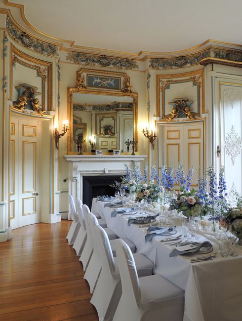 A long dining table set for a formal event in an elegant, historic room with ornate gold and white decor, intricate moldings, and large mirrors. The table is draped with a white tablecloth and decorat