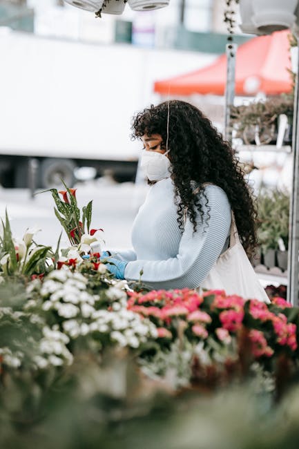 A woman with curly dark hair, wearing a white face mask and a light grey sweater, is selecting flowers at an outdoor flower stall. She is holding a flower arrangement in her gloved hands, examining th