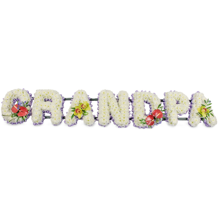 A personalised floral arrangement for a funeral or memorial service dominates the frame, the word "GRANDPA" carefully spelled in bold, upright letters that sit in a continuous line. Each letter is a small work of art in itself, densely packed with fresh white chrysanthemums so that no stems or foam are visible, only layers of soft petals forming a smooth, cushioned texture. The tribute is photographed against a clean white backdrop, allowing the refined details to stand out. Around the bottom of each letter, a slim edging of light lavender ribbon provides a subtle outline, its cool, soft hue echoing the calmness and introspection of a farewell in Highgate's peaceful surroundings. The letters are mounted on dark green connecting bars, which are mostly hidden but essential for keeping the piece straight and stable for display at the cemetery or crematorium. Four carefully placed floral sprays break up the white and introduce deeper meaning. On the "G" and on the "D", groups of rich red anthuriums with shiny, heart-shaped petals and protruding yellow spadices perch amidst glossy greenery and tiny flecks of purple filler flowers, adding intensity and passion to the tribute. On the first and final "A", sprays of lime-toned orchids unfold, their delicate petals and intricate markings exuding elegance and refined beauty. The interplay between the serene white chrysanthemums and the bolder accent flowers creates a flowing rhythm across the word, suggesting cherished memories threaded through grief. The tribute feels thoughtfully designed for families in Highgate and nearby Dartmouth Park who wish to honour a grandfather with something both traditional and quietly distinctive.